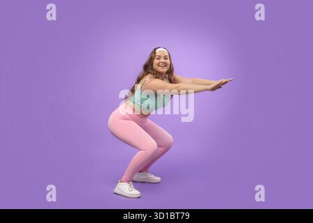 Hübsche junge Frau in Sportswear Squats auf lila Hintergrund für Fitness und Gesundheit Stockfoto