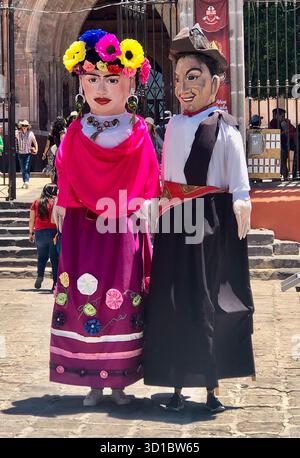 Riesige Marionettenfiguren, bekannt als Mojigangas, stehen vor einer Kirche während der farbenfrohen Los Locos Parade in San Miguel de Allende, Mexiko. Stockfoto