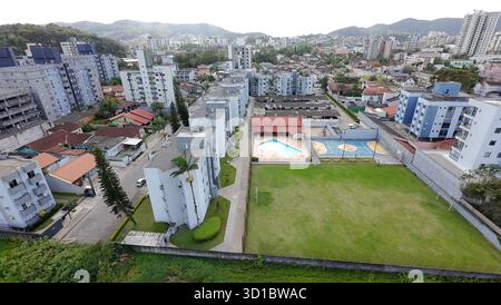 Aus der Vogelperspektive auf eine Wohngegend mit vielen Gebäuden, einem großen Swimmingpool, Fußball- und Futsal-Feldern, umgeben von Bäumen und Bergen unter einem Blau Stockfoto