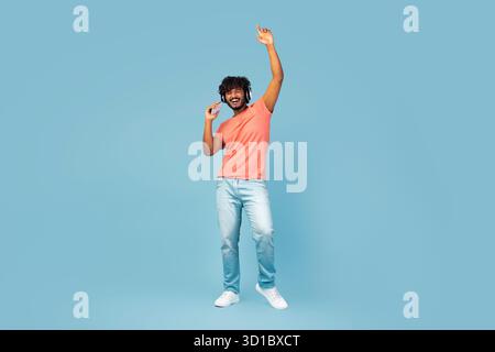 Junger Hindu-Typ, der Musik und Tanz in lässigem Outfit vor blauem Hintergrund genießt Stockfoto