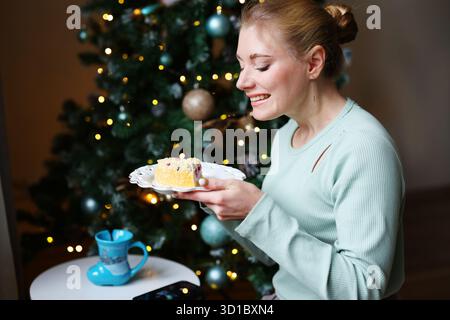 Smiling woman enjoying Christmas dessert and hot drink at home near decorated tree. Stockfoto