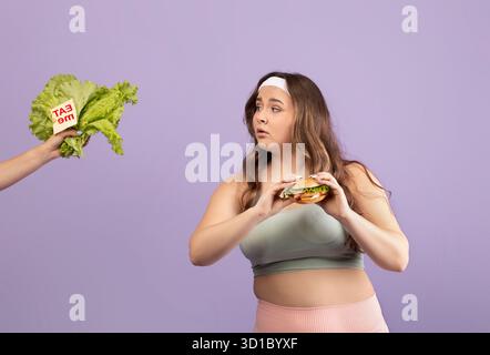 Die junge Frau in Sportswear wählt zwischen einem Burger und frischem Gemüse auf lila Hintergrund Stockfoto