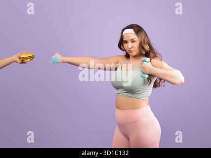 Pretty Plus Size Young Woman hebt Hantel, während sie einen Burger auf lila Hintergrund erhält Stockfoto