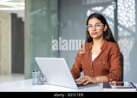 Indische Geschäftsfrau in Brille und Polka Dot-Bluse, die selbstbewusst auf einem Laptop an einem hellen, modernen Schreibtisch schreibt, konzentriert und professionell und blickt auf die Kamera Stockfoto