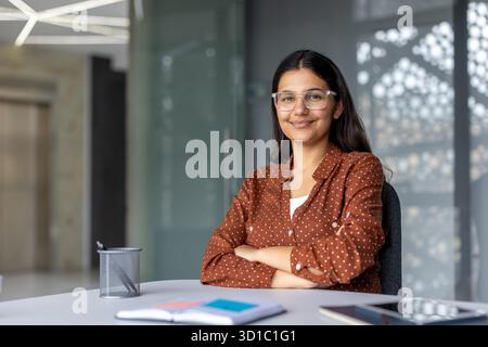 Junge indische Geschäftsfrau, die an einem Schreibtisch sitzt und selbstbewusst lächelt, während sie eine Brille und ein braunes Polka Dot-Shirt trägt. Sie vermittelt Professionalität und positive Einstellung Stockfoto