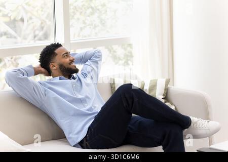 Sorgloser afrikanischer Kerl, der sich auf der Couch im gut beleuchteten Wohnzimmer entspannt Stockfoto