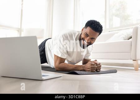 Ein lächelnder Afrikaner hat die Plankenposition, sieht auf den Laptop Stockfoto