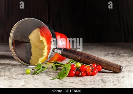 Eine Lupe liegt neben einem gebissenen roten Apfel und giftigen roten Beeren auf einer rustikalen Oberfläche. Konzeptuelles Stillleben symbolisiert detektiv investitig Stockfoto