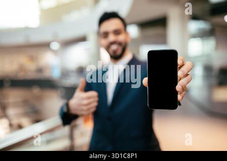 Geschäftsmann zeigt ein Smartphone mit leerem Bildschirm, während er im modernen Bürobereich die Daumen hochlegt Stockfoto