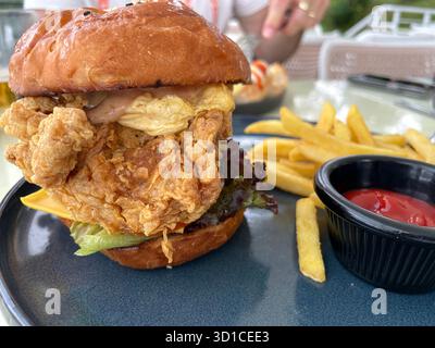 Nahaufnahme eines knusprigen gebratenen Hühnersandwiches mit Salat, Käse und Sauce, serviert mit Pommes frites und Ketchup auf einer dunkelblauen Keramikplatte Stockfoto