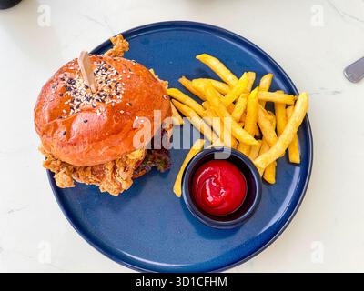Knuspriges, gebratenes Hähnchensandwich, serviert mit goldenen Pommes frites und Ketchup auf einer blauen Keramikplatte Stockfoto