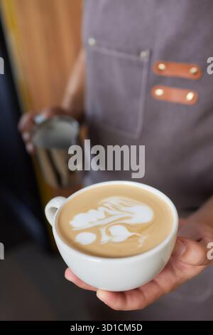 Bild von oben, in dem ein professioneller Barista Milch aus dem Becher in eine Tasse Kaffee eingießt, der von einem Barista zubereitet wird. Konzentrieren Sie sich auf die Hände, die eine Tasse halten Stockfoto