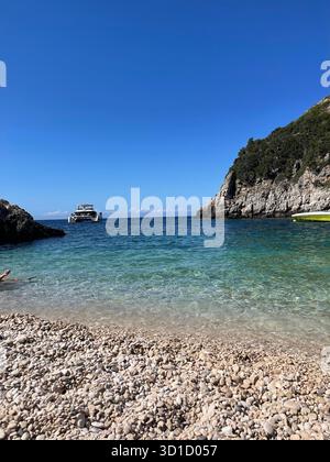 Idyllische abgeschiedene Pebble Beach Cove mit kristallklarem türkisfarbenem Wasser und felsigen, baumbedeckten Klippen unter einem tiefblauen Sommerhimmel mit einem Touristenboot Stockfoto