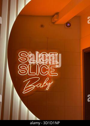 Warmes orangefarbenes Neonschild mit der Aufschrift „SLICE SLICE Baby“ an einer gekachelten Wand, das eine kreisförmige Leuchte in einer trendigen Pizzeria oder einem Restaurant reflektiert Stockfoto