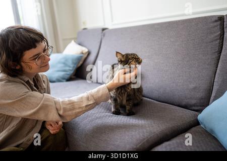 Junge, fröhliche Haustierbesitzerin, die Zeit zu Hause mit der Katze verbringt, neben dem Sofa sitzt und ihr Kätzchen streichelt Stockfoto