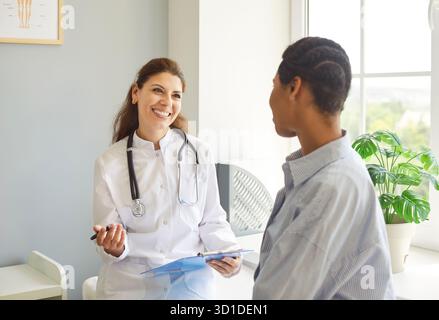 Ein glücklicher, lächelnder Arzt hält einen afroamerikanischen Patienten, der aufmerksam zuhört, beruhigend Stockfoto