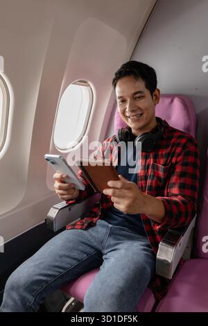 Ein junger Mann sitzt in einem Flugzeug und genießt seinen Flug, während er sein Smartphone und seinen Reisepass überprüft. Er wirkt entspannt und zufrieden und spiegelt die Moderne wider Stockfoto