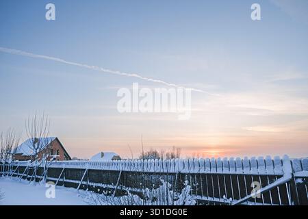 Wunderschöner Wintersonnenuntergang über verschneiten ländlichen Zaun und Haus auf dem Land. Friedliche Landschaft, Himmel mit Pastellfarben, frostiger Hof, ruhiger Abend. Perfec Stockfoto