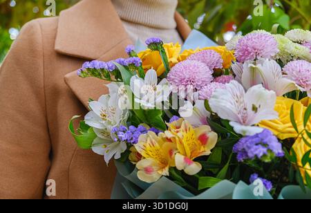 Frau im eleganten Mantel hält einen leuchtenden Blumenstrauß mit gelben, rosa, weißen und violetten Blüten. Perfekt für Feiern, Glückwünsche, Veranstaltungen, ca. Stockfoto
