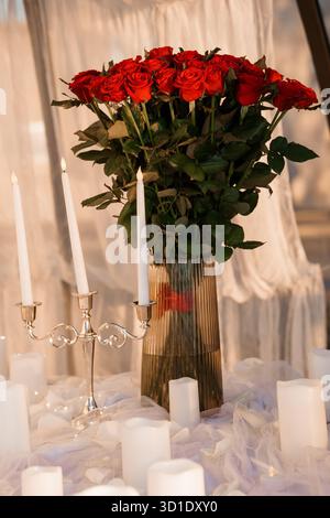 Eine atemberaubende Nahaufnahme fängt einen leuchtenden Blumenstrauß roter Rosen in einer strukturierten Glasvase zusammen mit beleuchteten weißen Kerzen ein, die an einen leidenschaftlichen Intim erinnern Stockfoto
