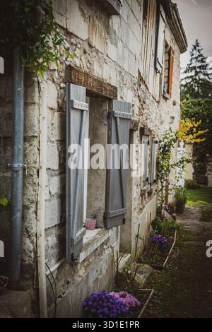 Ein bezauberndes Steingebäude mit verwitterten grauen Fensterläden und einem lebendigen Blumentopf verleiht der Szene einen Hauch von Farbe. Stockfoto