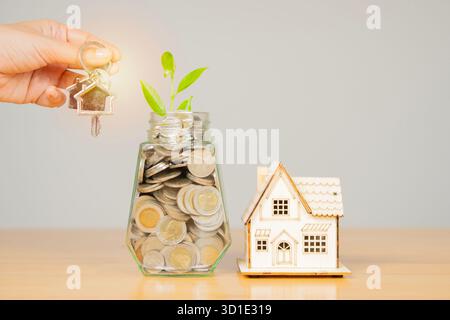 Hausschlüssel auf der Hand mit Geldgefäß Münzen und Hausmodell auf Holztisch. Investitions- und Immobilienkonzept, das Geld spart, um ein Haus zu kaufen Stockfoto