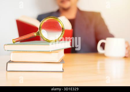 Nahaufnahme einer Lupe auf Büchern mit verschwommenem Bild eines sitzenden und lesenden Mannes. Selektiver Fokus. Stockfoto