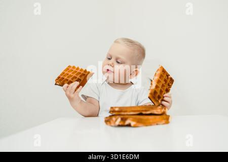 Ein niedliches Baby sitzt an einem Tisch und erkundet fröhlich eine bunte Auswahl an Waffeln, die pure Unschuld und Freude am Genuss köstlicher Leckereien zeigen Stockfoto