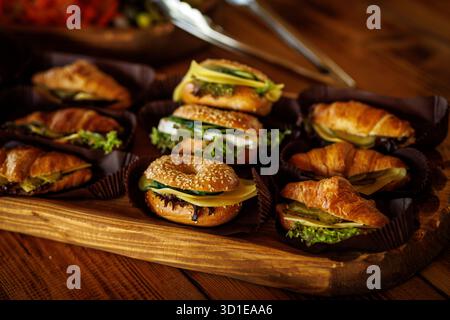 Holztablett mit frischen Mini-Sandwiches aus Croissants und Sesambagels gefüllt mit Käse, Salat und Gemüse. Stockfoto