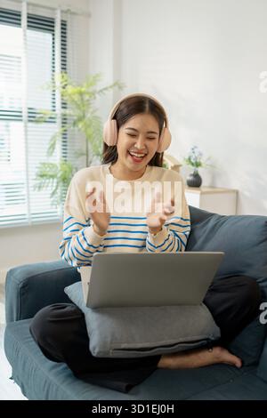 Eine junge Frau, die auf einem Sofa sitzt, Kopfhörer trägt, fröhlich lächelt, während sie vor ihren Laptop klatscht und während eines Onlins Freude und Aufregung zeigt Stockfoto