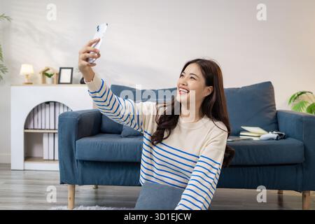Eine junge Frau macht mit ihrem Smartphone glücklich ein Selfie, während sie in einem gemütlichen Wohnzimmer auf dem Boden sitzt. Sie sieht fröhlich aus und schafft ein lässiges und Stockfoto