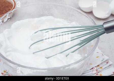 In einer klaren Schüssel können Sie geschlagenes Eiweiß aufbewahren, das für die Dessertzubereitung bereit ist. Daneben befindet sich eine Schüssel mit braunem Zucker und Eiern auf einer Blumenkerviette in einem hellen KI Stockfoto