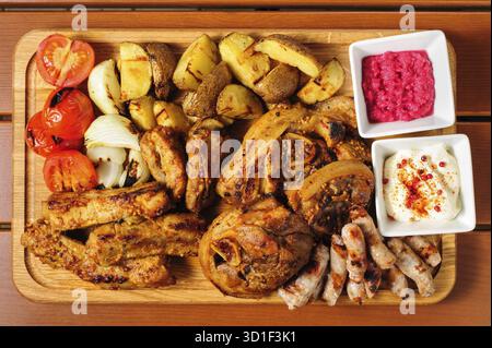 Großes Holzbrett mit Schweinerippchen und in Senfsauce gebackenen Steaks, kleinen Würstchen, gebackenen Kartoffeln, gegrillten Tomaten und Zwiebeln, heißer Sauce und Reiteraddis Stockfoto