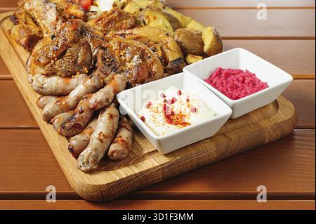 Großes Holzbrett mit Schweinerippchen und in Senfsauce gebackenen Steaks, kleinen Würstchen, gebackenen Kartoffeln, gegrillten Tomaten und Zwiebeln, heißer Sauce und Reiteraddis Stockfoto