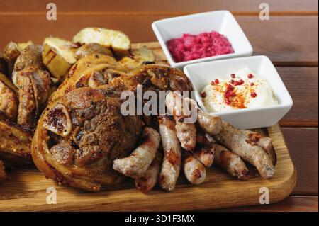 Großes Holzbrett mit Schweinerippchen und in Senfsauce gebackenen Steaks, kleinen Würstchen, gebackenen Kartoffeln, gegrillten Tomaten und Zwiebeln, heißer Sauce und Reiteraddis Stockfoto
