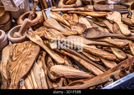 Handgefertigte Küchenartikel aus Olivenholz, darunter verschiedene Löffel, Spatel und Schneidebretter, füllen einen Verkaufsstand auf einem lebhaften Markt und präsentieren natürliche PA Stockfoto