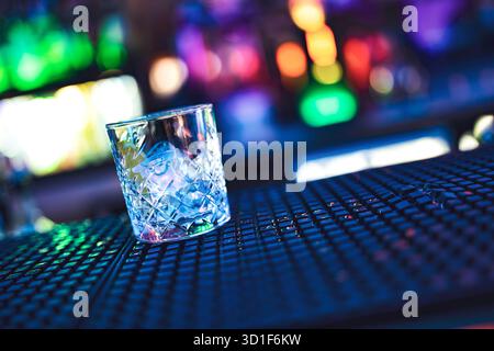 Kristallglas gefüllt mit klaren Eiswürfeln auf einer strukturierten Bartheke, beleuchtet von lebendigen, unscharfen Neonlichtern, die eine dynamische Nacht schaffen Stockfoto