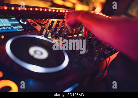 DJ-Hände passen die Bedienelemente am professionellen Mixer und Plattenspieler an, mischen elektronische Titel und gestalten den Sound für eine lebendige Nachtclub-Party mit Licht Stockfoto