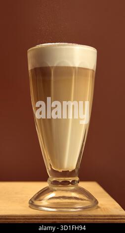 Latte Macchiato in hohem Glas mit mehrschichtigem Milchschaum und bestreuseltem Kakaopulver auf braunem Hintergrund Stockfoto