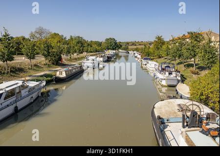 Steg am Canal du Midi, hier ein langsam-brauner Wasserweg, mit verankerten Hausbooten und Motoryachten auf beiden Seiten, gerahmten Uferpflanzen und einigen Bauten Stockfoto