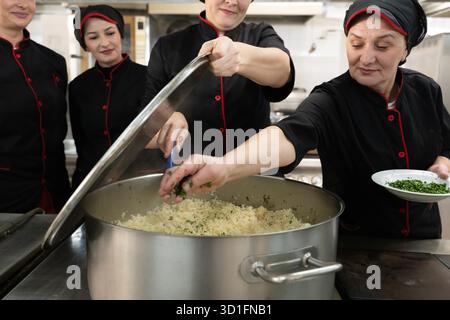 Eine Gruppe von Köchen in schwarzen Uniformen bereitet Reis in einem großen Topf in einer kommerziellen Küche zu. Weibliche Köchinnen rühren und fügen Kräuter hinzu, während sie als Team arbeiten. Konzept für Teamwork, Gastronomie und Gastgewerbe || Modell veröffentlicht Stockfoto