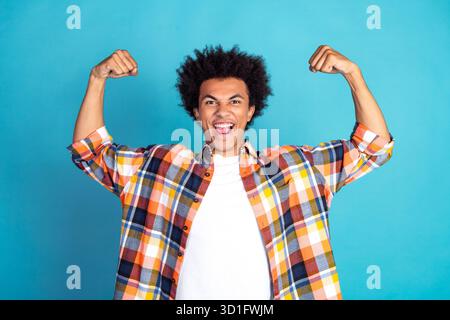 Junger Mann in kariertem Hemd, der mit Freude und Zuversicht die Muskeln vor blauem Hintergrund spannt Stockfoto