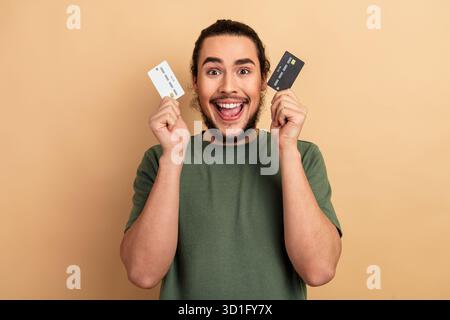 Das junge männliche Model hält zwei Kreditkarten und lächelt mit grünem T-Shirt vor beigefarbenem Hintergrund für ein Lifestyle-Fashion-Shopping-Promo Stockfoto