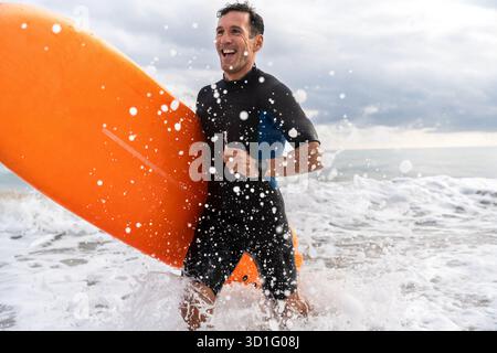 Begeisterter Mann in einem Neoprenanzug mit einem orangen, weichen Surfbrett, stehend im flachen Meereswasser mit Wellen, die plätschern, lachen und das e genießen Stockfoto
