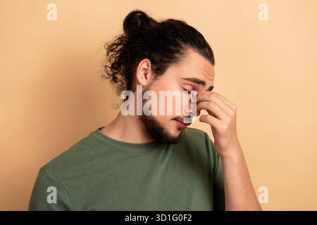 Junger männlicher Mixed-Race-Model mit langen Haaren und Bart, der ein grünes T-Shirt trägt, das Stress und Erschöpfung vor einem beigefarbenen Hintergrund ausdrückt Stockfoto