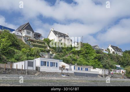 Eine Reihe von farbenfrohen, historischen Strandhütten mit Wänden aus Naturstein säumen den felsigen Strand am Fuße eines steilen Hanges, auf dem Villen thronen Stockfoto