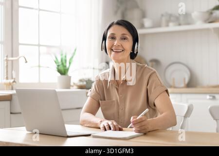 Lächelnde reife Frau, die online am Notebook sitzt und in Kopfhörern sitzt Stockfoto