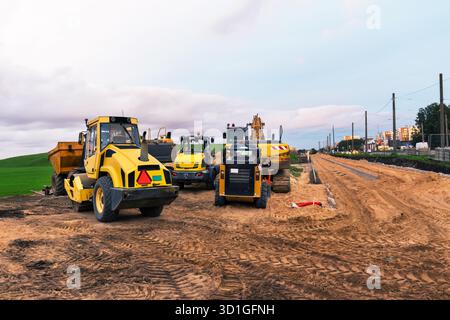 Straßenbauszene mit einer Straßenwalze, einem Bagger, einem Minikipper und einem Traktor auf einer unbefestigten Straße. Schwere Maschinen werden auf dem Standort mit einer Wolke geparkt Stockfoto