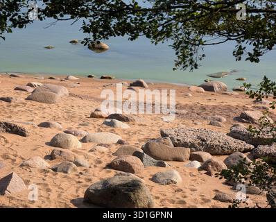Eine ruhige Strandszene mit glatten Felsen und goldenem Sand, mit ruhigem Wasser und sanften Wellen, umgeben von üppigem Grün. Stockfoto