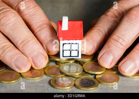 Konzeptionelles Bild, das Hände zeigt, die ein kleines Hausmodell auf Euro-Münzen schützen, das Eigenheimersparnis, Immobilieninvestitionen und finanzielle Sicherheit repräsentiert Stockfoto